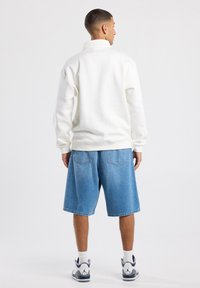 Felpa bianca con colletto alto, abbinata a pantaloni shorts in denim azzurro chiaro. Il modello indossa sneakers bianche con dettagli grigi e calzini alla caviglia.