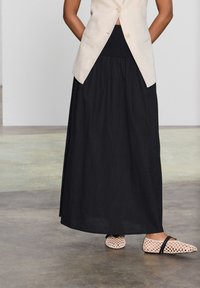 Jupe noire, longue jusqu'au sol, en tissu léger, associée à un blazer en lin beige et des ballerines roses tissées avec des accents noirs.