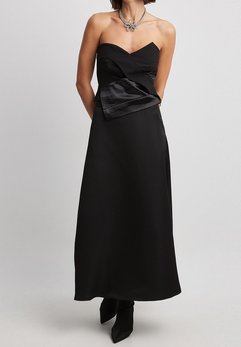 Robe noire sans bretelles avec un corsage ajusté et des détails en satin drapé à la taille. Longueur au sol, texture lisse, associée à des bottines noires.