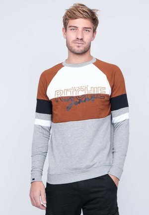 Jeune homme aux cheveux châtain clair portant un sweat-shirt à manches longues color-block avec le texte "RITCHIE", debout devant un fond clair uni.