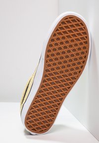 Semelle d'une basket avec un motif de crampons couleur brun waffle, une semelle intermédiaire blanche et une tige texturée jaune, inclinée contre une surface blanche.