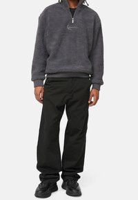 Grå fleecepullover med halvlukning, der har en tekstureret overflade og lille broderet detalje. Kombineret med løstsiddende sorte bukser og sorte sko.