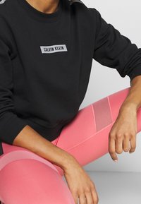 Sweatshirt noir à col rond avec un patch logo "CALVIN KLEIN" gris, associé à des leggings roses avec des détails en mesh et des rayures.