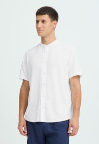 Chemise blanche en lin à manches courtes avec un col mandarin, dotée d'une fermeture boutonnée à l'avant et d'une coupe décontractée. Texture lisse, design simple.