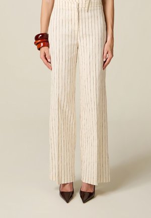 Vrouw die roomkleurige wijde broek met dunne verticale strepen draagt, zwarte puntige schoenen en dikke bruine armbanden aan haar linkerpols.