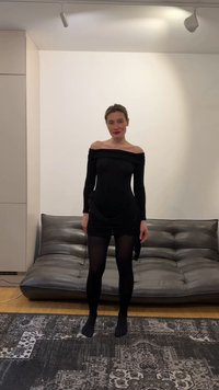 Vrouw in zwarte off-shoulder jurk en panty staat blootsvoets voor een grijze leren bank op een patroonrijke vloerkleed in de woonkamer.