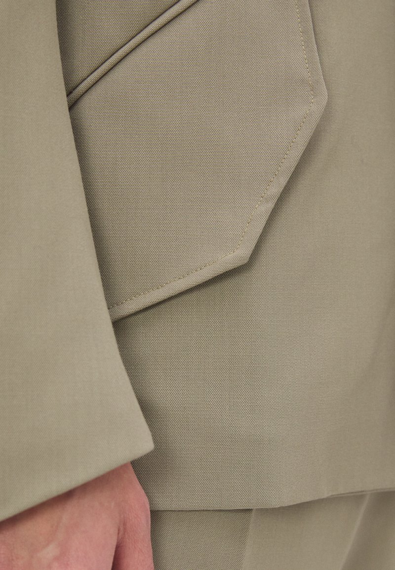 Beiger Stoff mit glatter Textur, der ein geometrisches Taschendetail und kontrastierende Nähte aufweist. Das Design umfasst scharfe Linien und eine strukturierte Form.