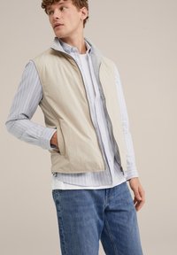 Beige vest met een grijze kraag, gecombineerd met een blauw-wit gestreepte overhemd met knopensluiting. Casual blauwe spijkerbroek maakt de outfit compleet.