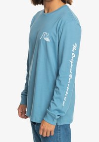 Light blue long-sleeve t-shirt made of cotton, featuring a small wave graphic on the chest and white text on the right sleeve.