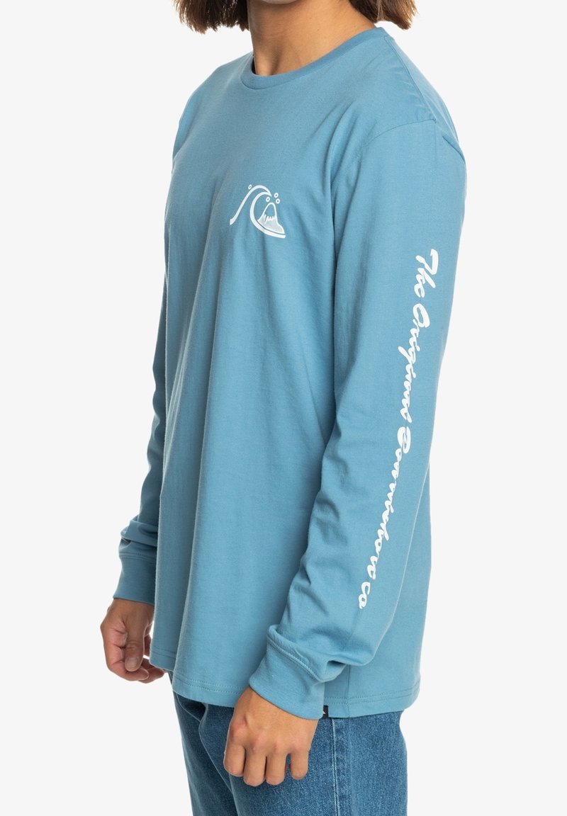 Light blue long-sleeve t-shirt made of cotton, featuring a small wave graphic on the chest and white text on the right sleeve.