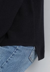 Marineblauer Strickpullover mit strukturiertem Muster und seitlichem Schlitz. Kombiniert mit hellblauen Jeans aus Denim, die einen lässigen Look präsentieren.