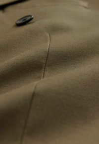 Olive zaļas auduma tuvplāns, kuram ir gluda tekstūra, viegla spīduma un apaļa melna poga ar šuvju detaļām.