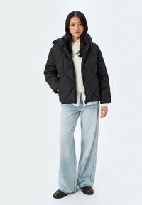 Sort puffjakke med diamant-quilt mønster, tætsiddende hætte og lynlås lukning, parret med løse lyseblå jeans og mørke sko.