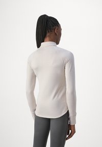 Haut long à manches beige clair avec une texture ribbée subtile, col haut et ourlet arrondi, associé à des leggings ajustés gris foncé.