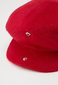 Rode wollen nieuwsjongenspet met metalen drukknoopdetails op de voor- en zijpanelen, weergegeven op een lichte achtergrond.
