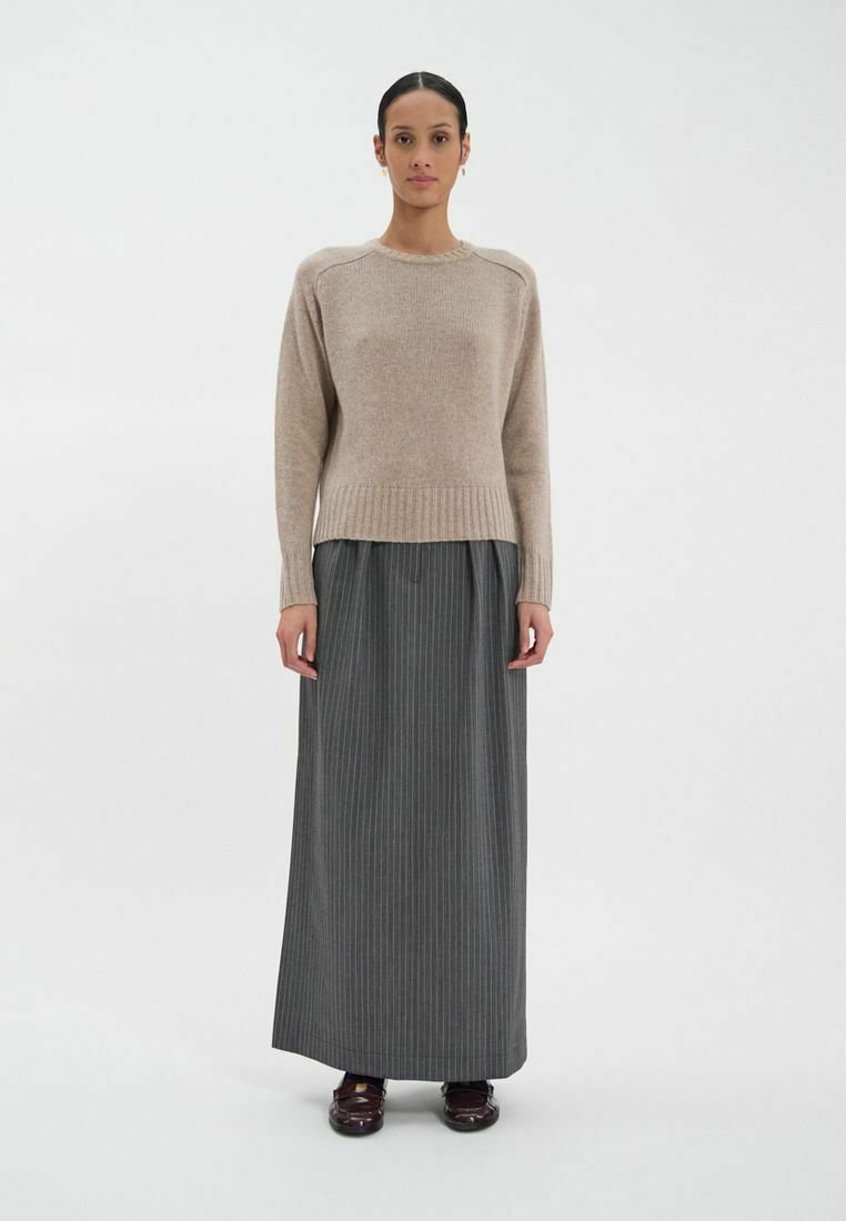 Un pull en tricot beige avec un col arrondi, associé à une longue jupe gris à rayures. La tenue présente une coupe décontractée et des poignets côtelés.