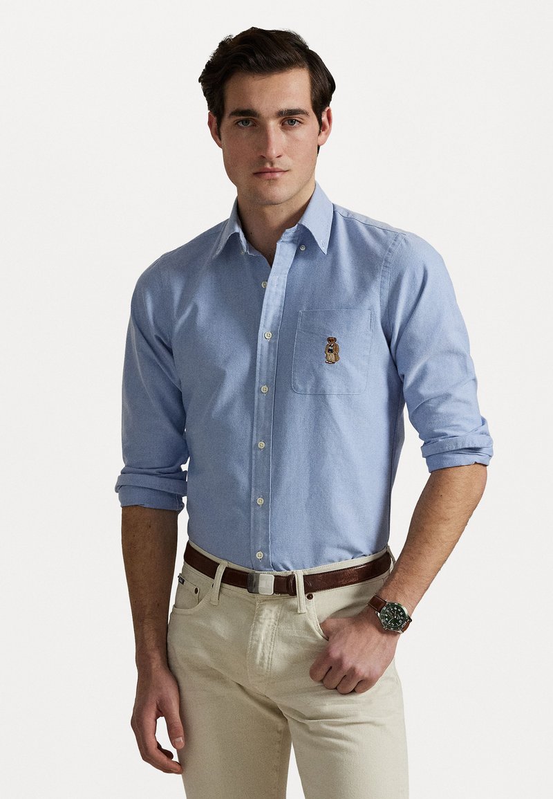 Chemise bleu clair à manches longues avec une poche poitrine arborant un logo brodé. Manches retroussées et col classique, associée à un pantalon beige et une ceinture marron.