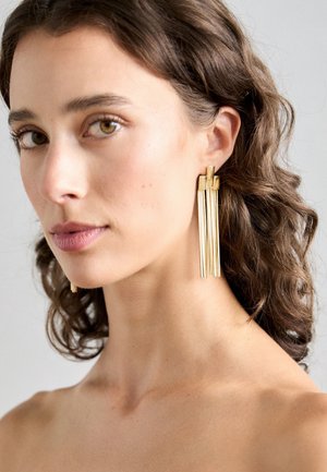 Mujer con cabello castaño corto y rizado, que lleva pendientes largos de barra dorada con flecos, mirando ligeramente hacia la izquierda sobre un fondo gris claro.