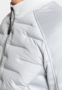 Hellegraue gepolsterte Jacke mit hohem Kragen, Reißverschluss und kontrastierenden, glatten Stoffakzenten auf den Schultern. Strukturiertes Oberflächenbild.