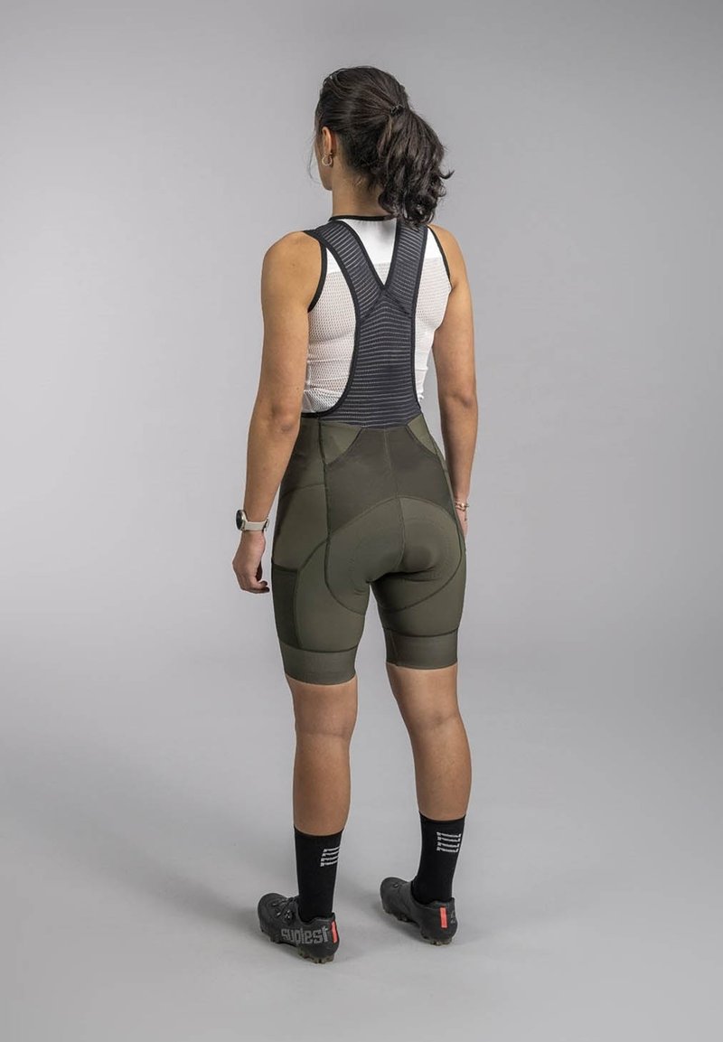 Bib shorts in dark green fabric with padded rear, featuring a black mesh upper and fitted design. Worn with cycling shoes and socks.