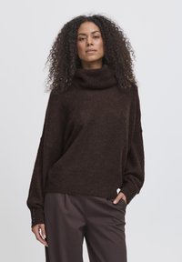 Maglione over-size marrone con collo a imbuto, tessuto texturizzato e maniche lunghe. Presenta dettagli a bottone sui polsini, abbinato a pantaloni scuri.