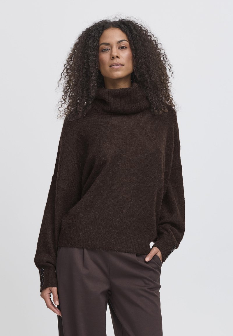 Maglione over-size marrone con collo a imbuto, tessuto texturizzato e maniche lunghe. Presenta dettagli a bottone sui polsini, abbinato a pantaloni scuri.