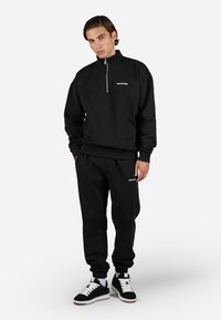 Black sweatshirt with a half-zip, ribbed cuffs, and a logo. Paired with matching black sweatpants and black sneakers with white accents.