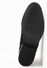 Zapato de ante negro con suela de goma lisa y patrón texturizado, que presenta una etiqueta grabada con "RIVER ISLAND" y detalles de tamaño.