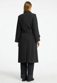 Črn plašč v ribji kost, z pasom, širokimi reverji in gumbnimi manšetami, v kombinaciji s temnimi hlačami in ravnimi čevlji.