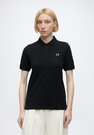 Femme aux cheveux blonds courts portant un polo noir avec un petit logo brodé sur la poitrine et un pantalon blanc, debout devant un fond blanc.