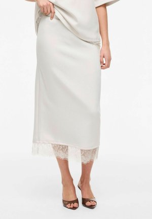 Vrouw draagt een witte satijnen midirok met kanten rand aan de zoom, gecombineerd met bruine open-toe hakmuiltjes tegen een effen achtergrond.