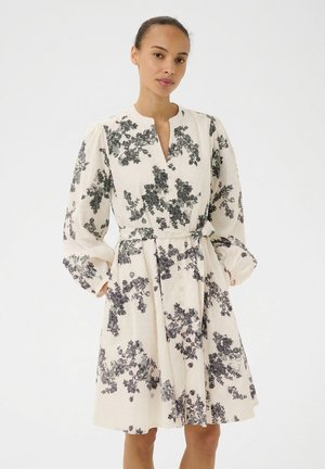 Kobieta z włosami związanymi do tyłu, ubrana w białą sukienkę za kolano z długimi rękawami, czarnym kwiatowym wzorem i paskiem wiązanym w talii.