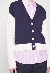 Marinblå och krämfärgad kofta med V-ringning, fem knappar och ribbade ärmslut, lagd över en rosa och vit randig skjorta.