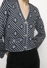Cárdigan de estampado negro y azul que presenta un profundo escote en V, cierre de botones, dos bolsillos frontales y un diseño de rayas audaces con formas geométricas.