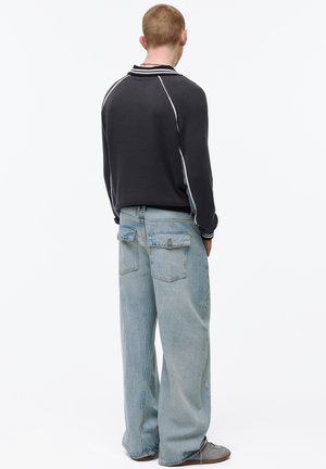 Muž v černém tričku s dlouhým rukávem a bílými pruhy a volných světle modrých džínách stojí zády k divákovi na bílém pozadí.