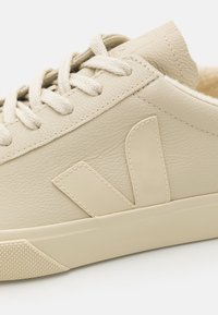 Detailní záběr béžové kožené tenisky s odpovídajícími tkaničkami a vyšitým logem "V" na boku, která má strukturovanou gumovou podrážku.