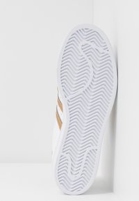 Biała podeszwa do trampków z teksturowanymi rowkami i falistym wzorem, z brązowymi i złotymi paskami po bokach oraz gumową konstrukcją.