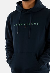Marineblauwe hoodie met een kangoeroe-pocket, voorzien van een groen logo "TOMMYJEANS" en een klein rood-wit embleem aan de linkerkant. Zacht, katoenmix stof.