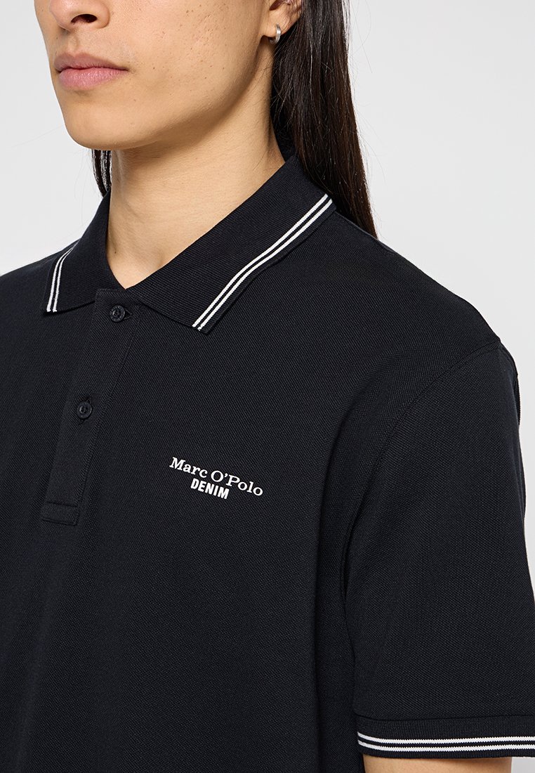 Schwarzes Poloshirt aus strukturiertem Material. Verfügt über einen klassischen Kragen mit weißen Streifen und ein weißes Logo auf der linken Brustseite.