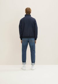 Chaqueta bomber de color azul marino con un cuello alto, combinada con unos vaqueros de denim ligeros y puños doblados. Costuras naranjas en los vaqueros, zapatillas blancas.
