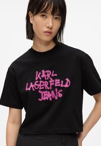 Černé bavlněné tričko s kratším střihem. Na přední straně se výrazně nachází růžový nápis „KARL LAGERFELD JEANS“ nastříkaný sprejem.