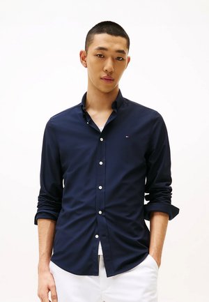 Camicia blu marina con bottoni, realizzata in tessuto liscio, con maniche lunghe arrotolate ai polsini e un piccolo logo sul petto. Bottoni bianchi.