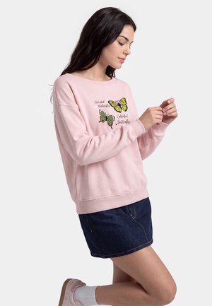 Donna con felpa rosa con illustrazioni di farfalle e testo, gonna in denim scuro, calzini bianchi e scarpe rosa chiaro, che posa con un ginocchio a terra.