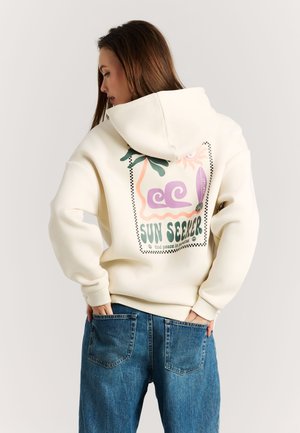 Vrouw draagt een crèmekleurige hoodie met een kleurrijke "Sun Seeker" afbeelding en tekst op de achterkant, gecombineerd met een blauwe spijkerbroek, handen in de achterzakken.