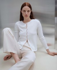 Weiße strukturierte Strickjacke mit runder Halslinie, langen Ärmeln und sechs Knöpfen, kombiniert mit hellbeigen weiten Hosen.