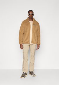 Lichte fleece jack in tan met rits en logopatch, gecombineerd met een lichtbeige t-shirt, cargobroek en grijze wandelschoenen.