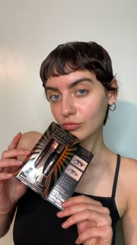 Schwarze Mascara-Verpackung mit goldenen Akzenten, die Vorher-Nachher-Bilder der Augen zeigt. Die Halterin posiert mit glattem Haar und minimalem Make-up.