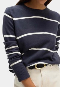 Granatowy sweter w białe paski, wykonany z dzianiny. Posiada okrągły dekolt i prążkowane mankiety. Połączony z kremowymi spodniami i czarnym paskiem.