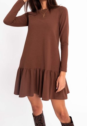 Vrouw draagt een bruine jurk met lange mouwen en een gerimpelde zoom, gecombineerd met knielaarzen met slangenhuidpatroon en een gouden kruisje aan een ketting.