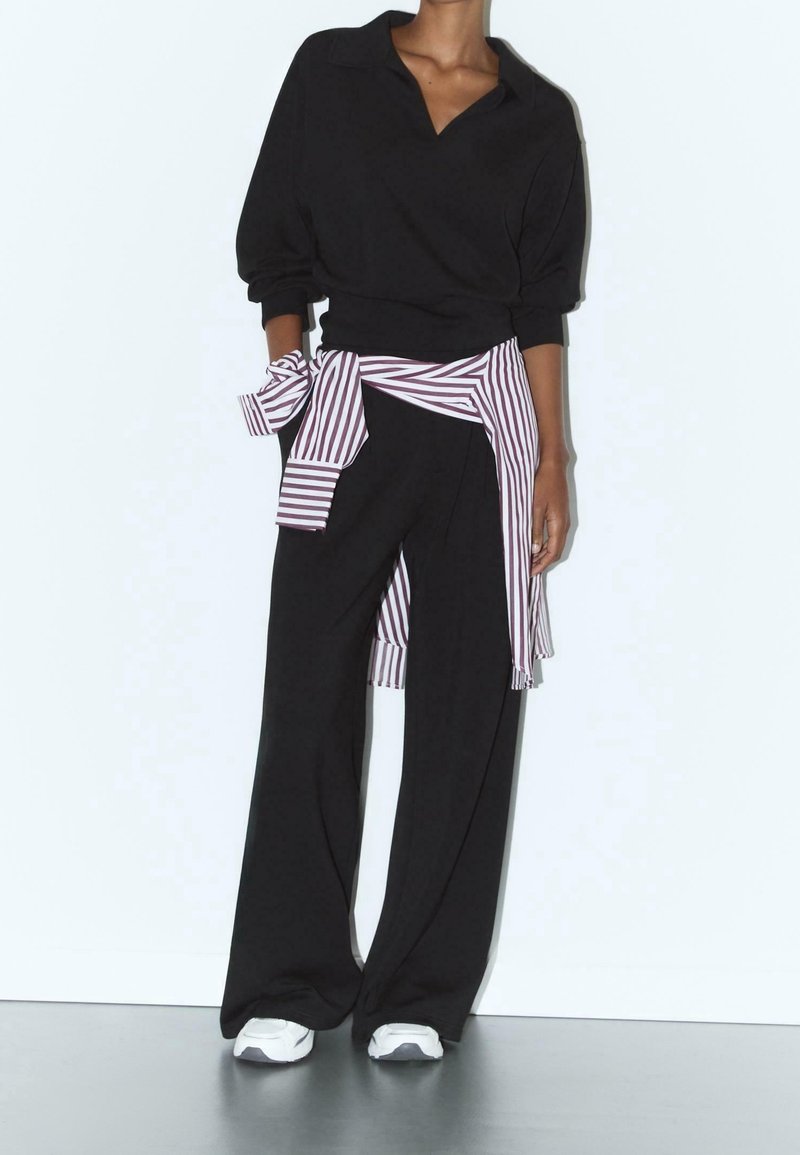 Personne portant un pantalon noir ample, un haut noir à manches longues avec col, des baskets blanches et une chemise rayée nouée autour de la taille.